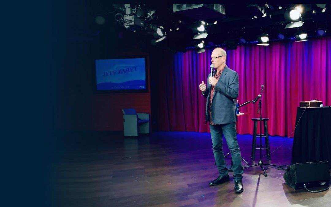 Jeff Zaret stands on stage, speaking into a microphone with red curtains and stage lights behind him.