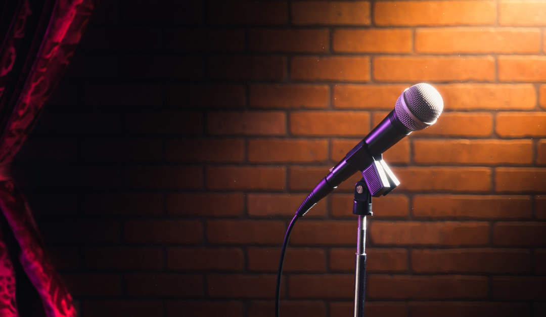 A microphone on a stand is lit by a spotlight against a brick wall, with a red curtain to the side, setting the perfect stage for stand up comedy.