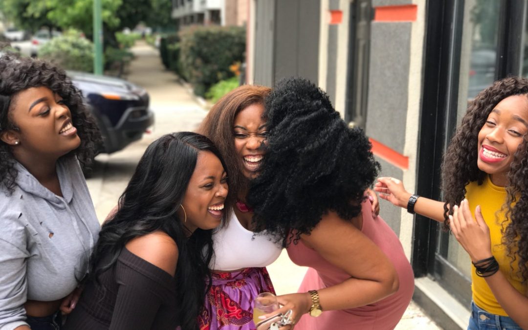 Five women laughing and smiling together outside on a sidewalk, sharing a casual, happy moment and showcasing their natural comedy skills.