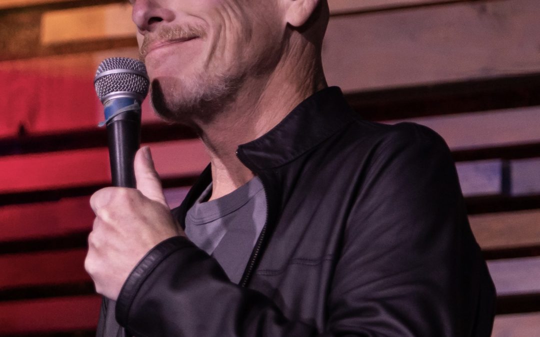 A man in a black jacket smiles while holding a microphone on stage with wooden panels behind him.