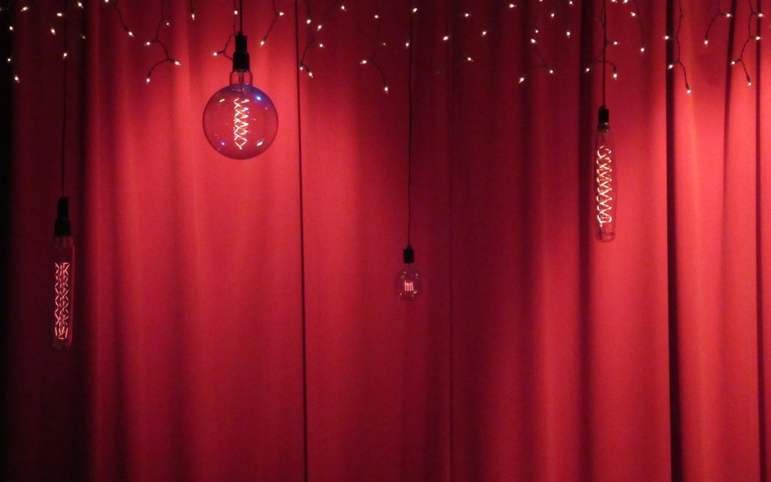 Hanging lightbulbs and string lights glow against deep red curtains, setting the scene for a touch of stage fright.