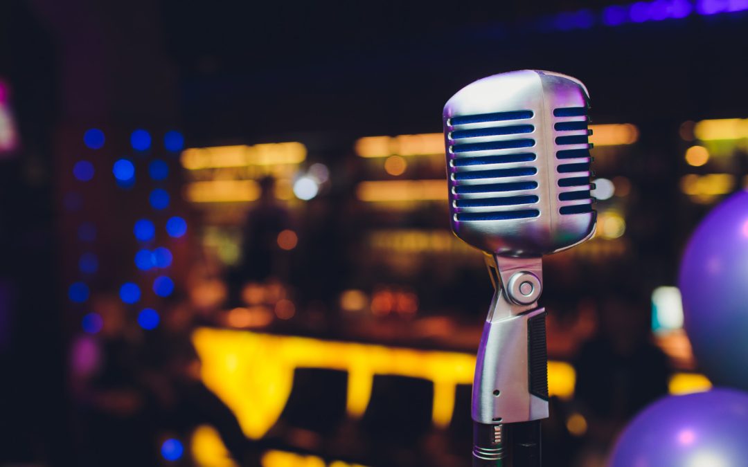 A vintage silver microphone stands in focus with colorful lights and a blurred bar background, ready to showcase humor's healing power.